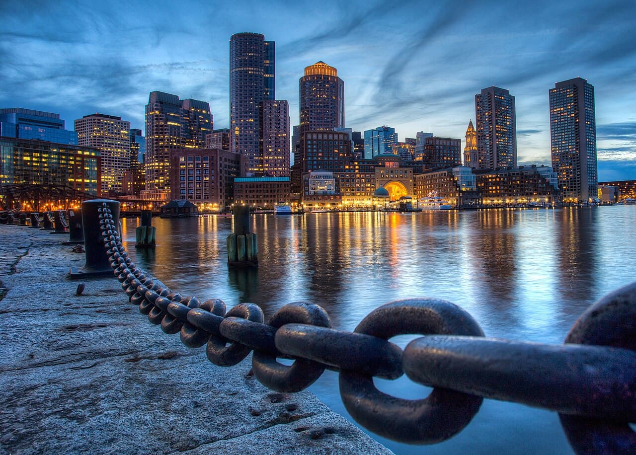 Downtown Boston's towers and the Rowes Wharf arch lit at dusk, seen across the harbor from Fan Pier in the Seaport with a heavy chain in front