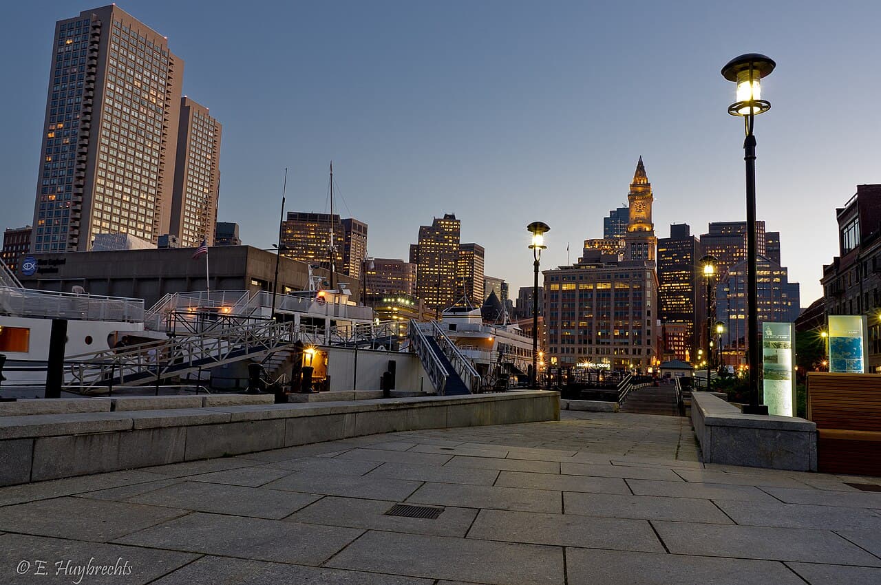 The paved walkway on Long Wharf in Boston at dusk, with docked boats on the left and the Custom House Tower lit up ahead