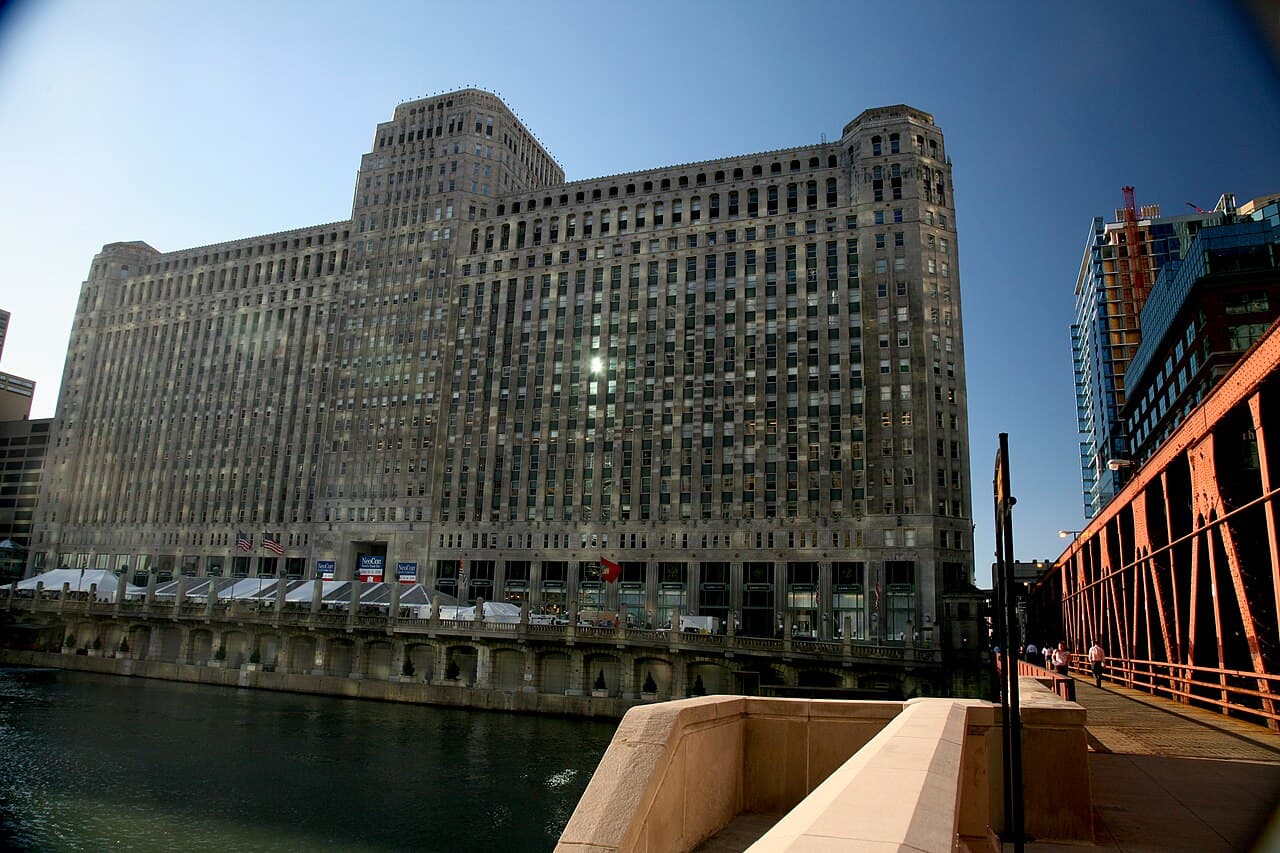 The Merchandise Mart, a huge stone building, seen across the Chicago River from a red steel bridge on a clear day