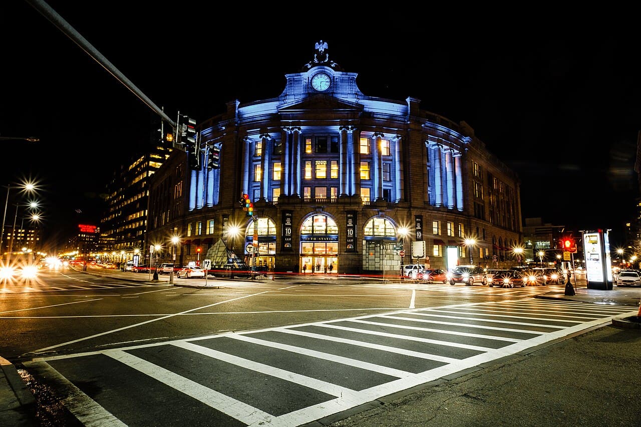 South Station's stone headhouse in Boston lit blue at night, with its clock and eagle above the arched entrance and a crosswalk in front