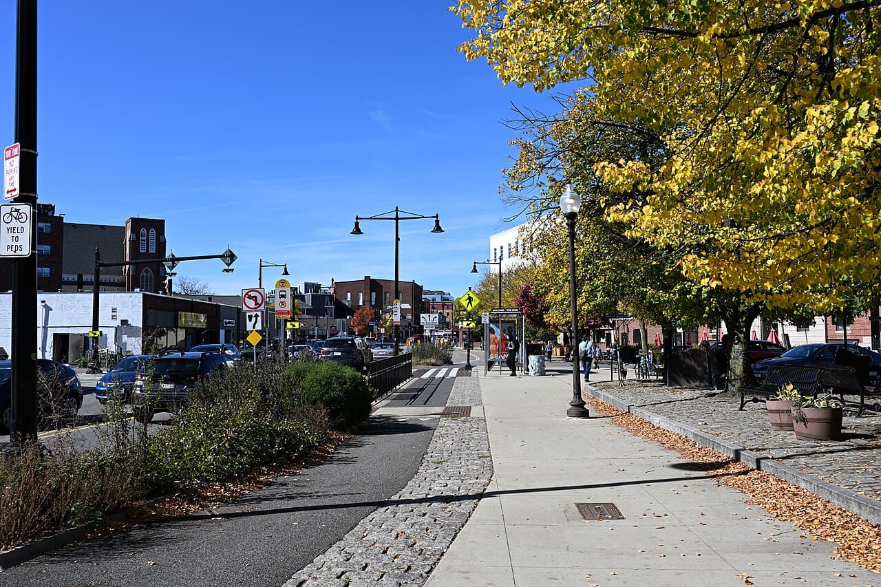 A sidewalk and bike lane through Union Square in Somerville under yellow autumn trees, with low brick buildings ahead