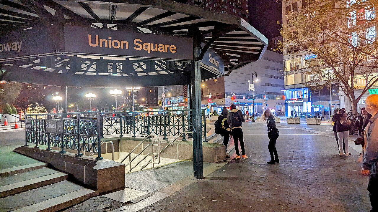 A lit Union Square subway entrance, with a canopy over the stairs and people standing beside it at night