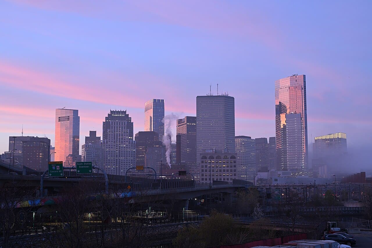 The downtown Boston skyline under a pink and purple sky, seen from West Broadway in South Boston with the highway in front