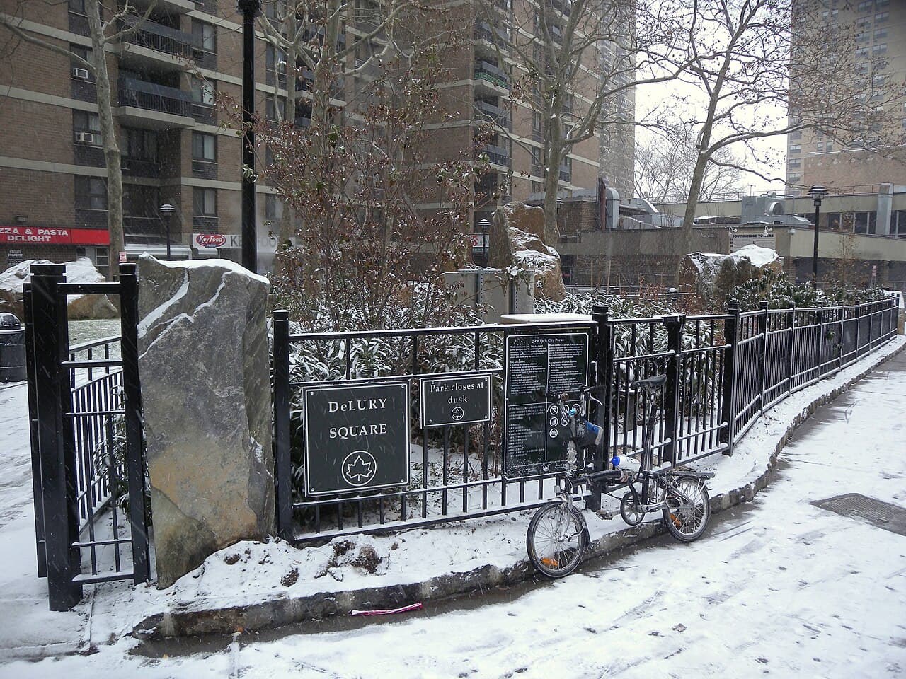 DeLury Square at Fulton and Gold Streets under snow, with a bicycle against its fence