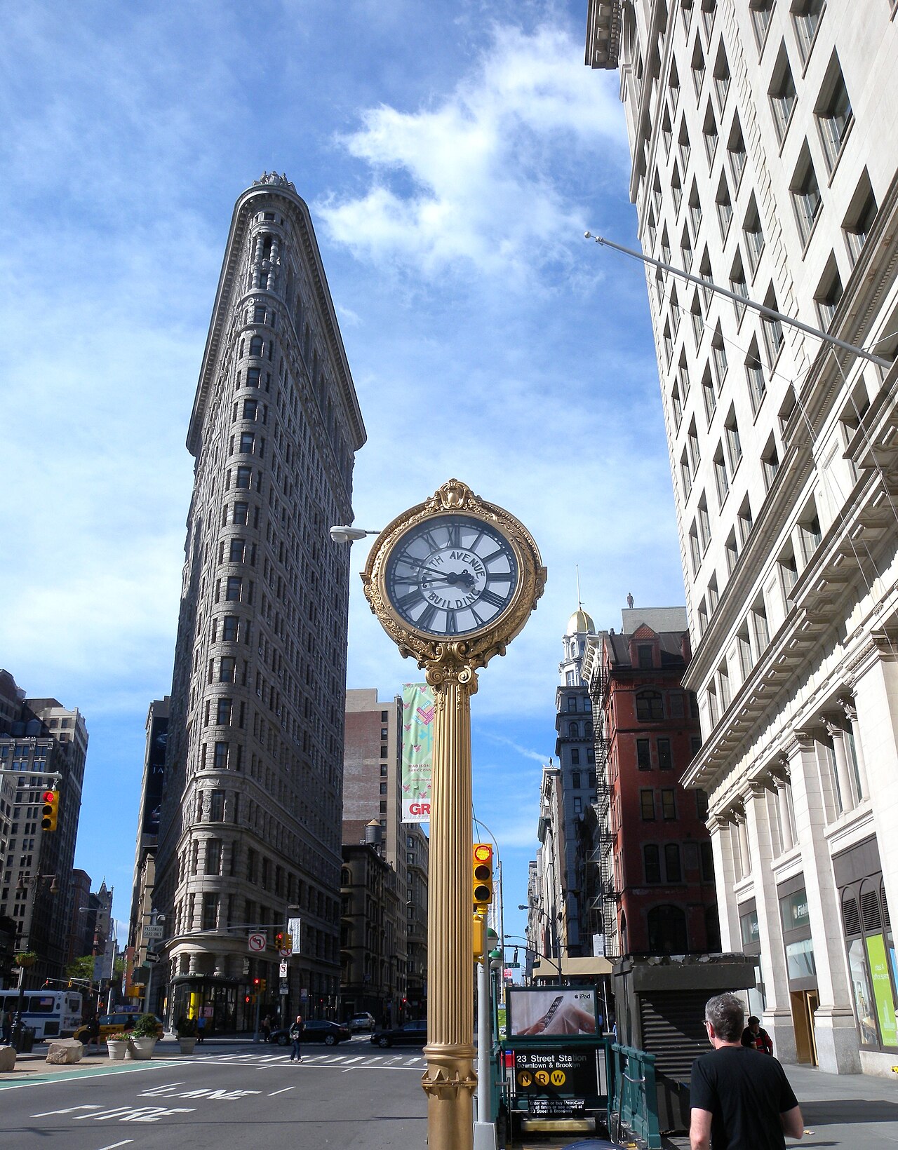 The Flatiron Building and the gold sidewalk clock at 200 Fifth Avenue on a sunny day, with a subway entrance beside the clock