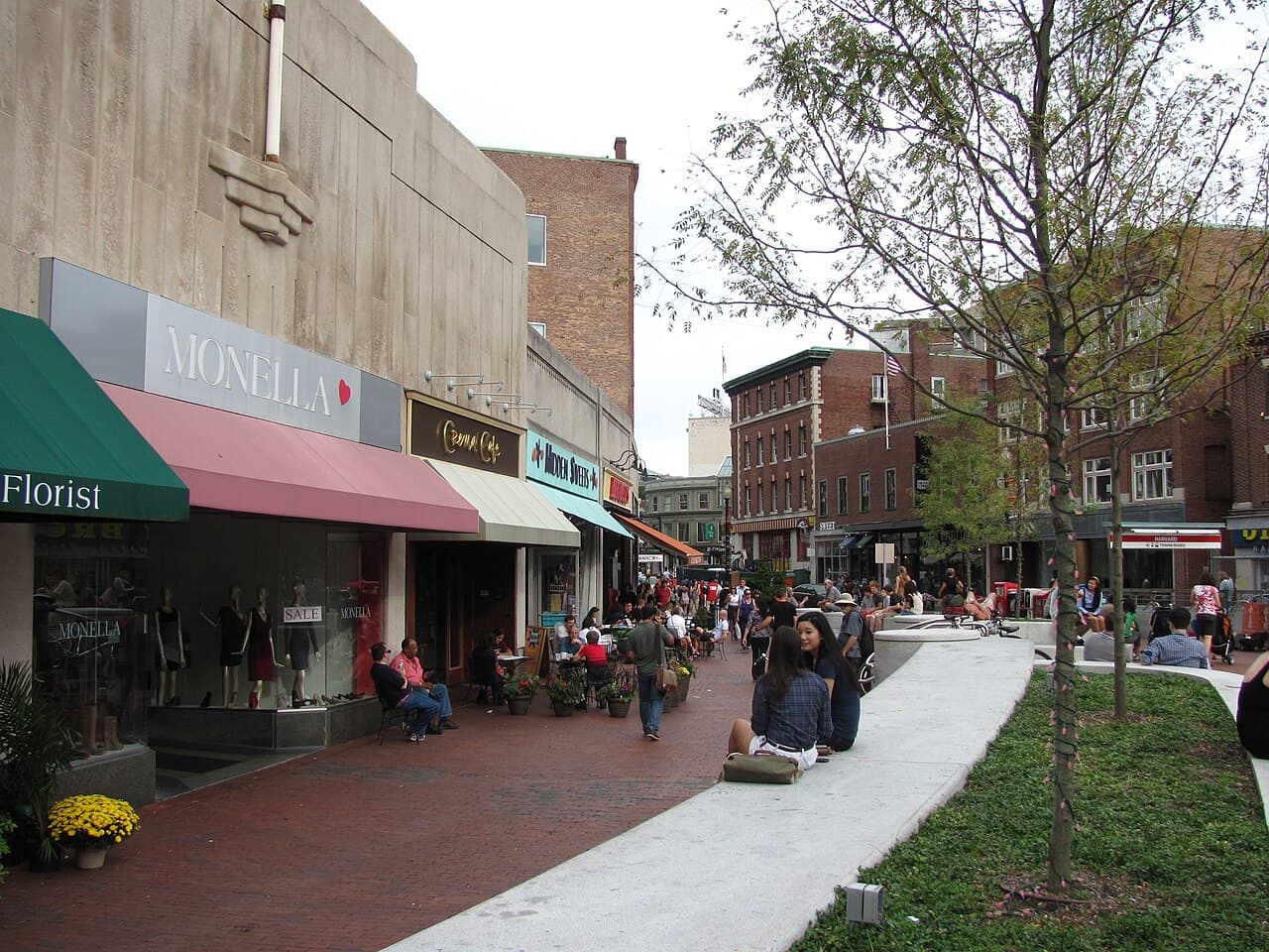 People walking and sitting along a brick sidewalk lined with shops in Harvard Square, Cambridge