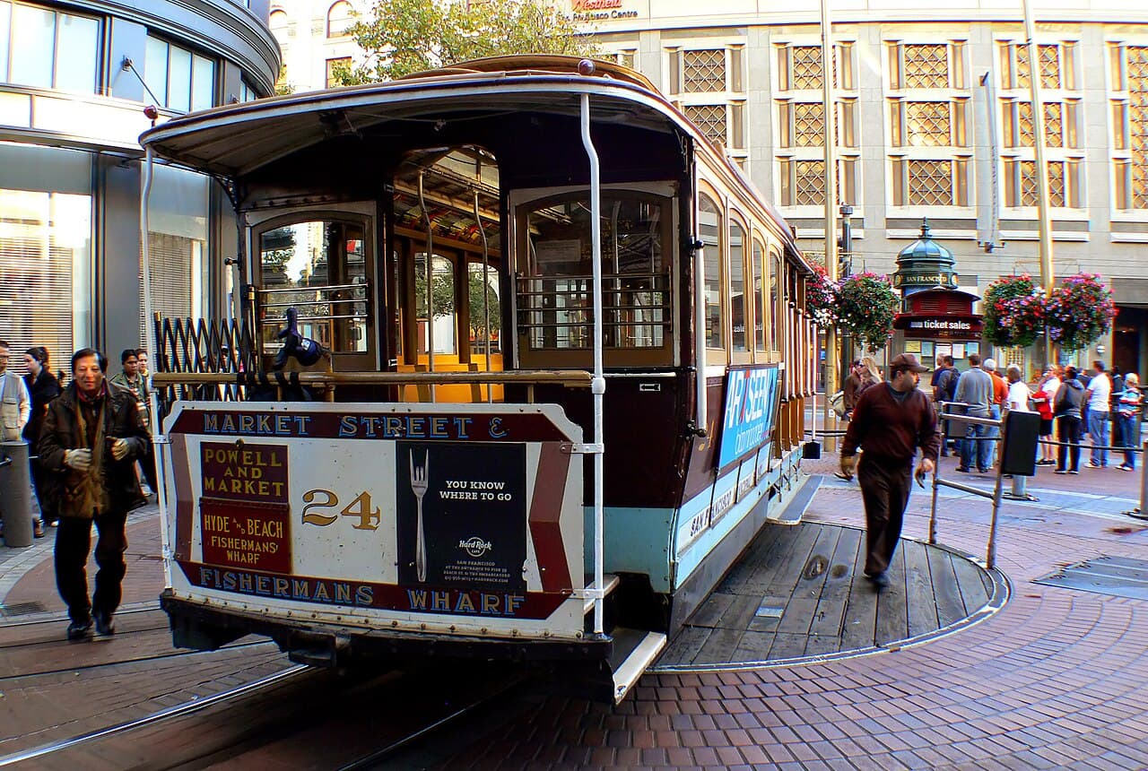 A cable car on the turntable at Powell and Market Streets, with people walking past and shops behind it