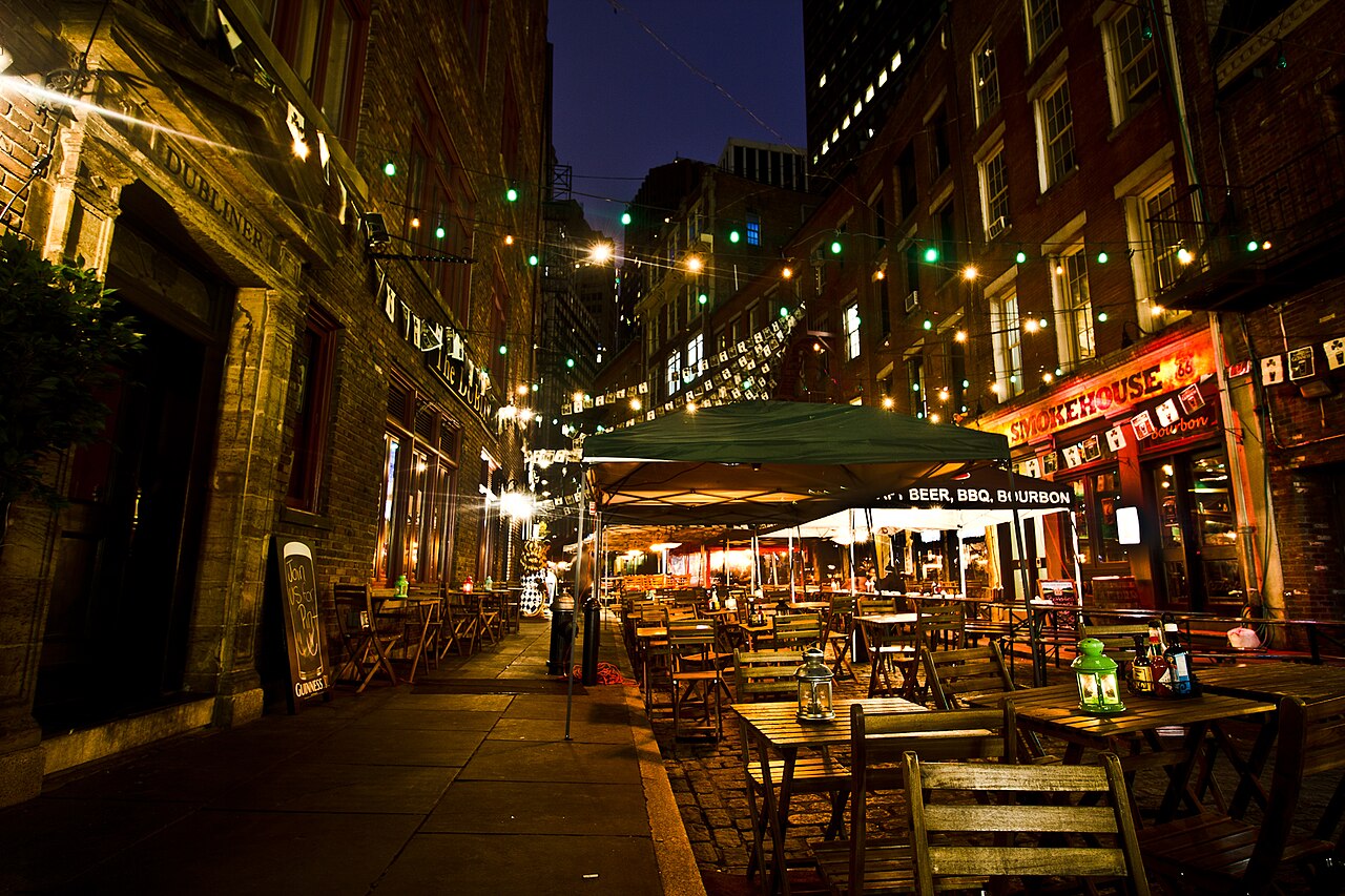 Outdoor tables under a tent and string lights on Stone Street, with lit restaurant fronts on both sides
