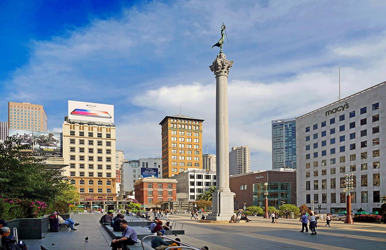 People sitting on the steps of Union Square's plaza in San Francisco, with the Dewey Monument column and department stores around it